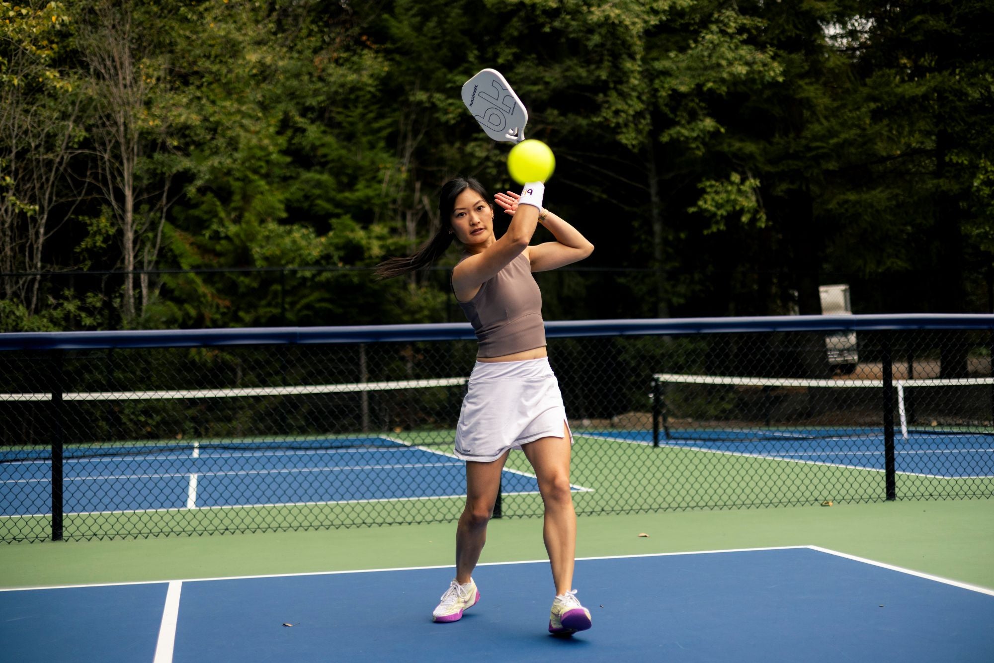 Woman playing pickleball on a court with trees in the background