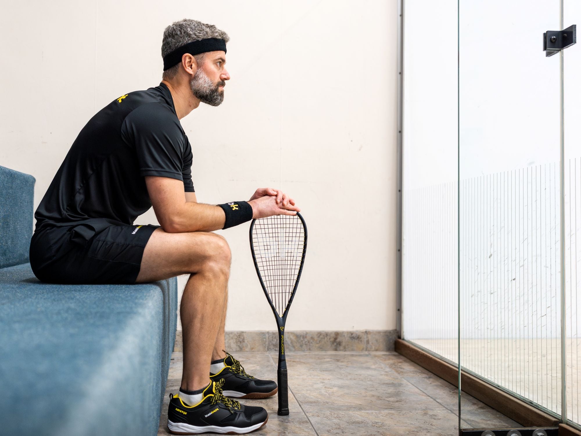 Man sitting on a bench holding a squash racket looking into a squash court.