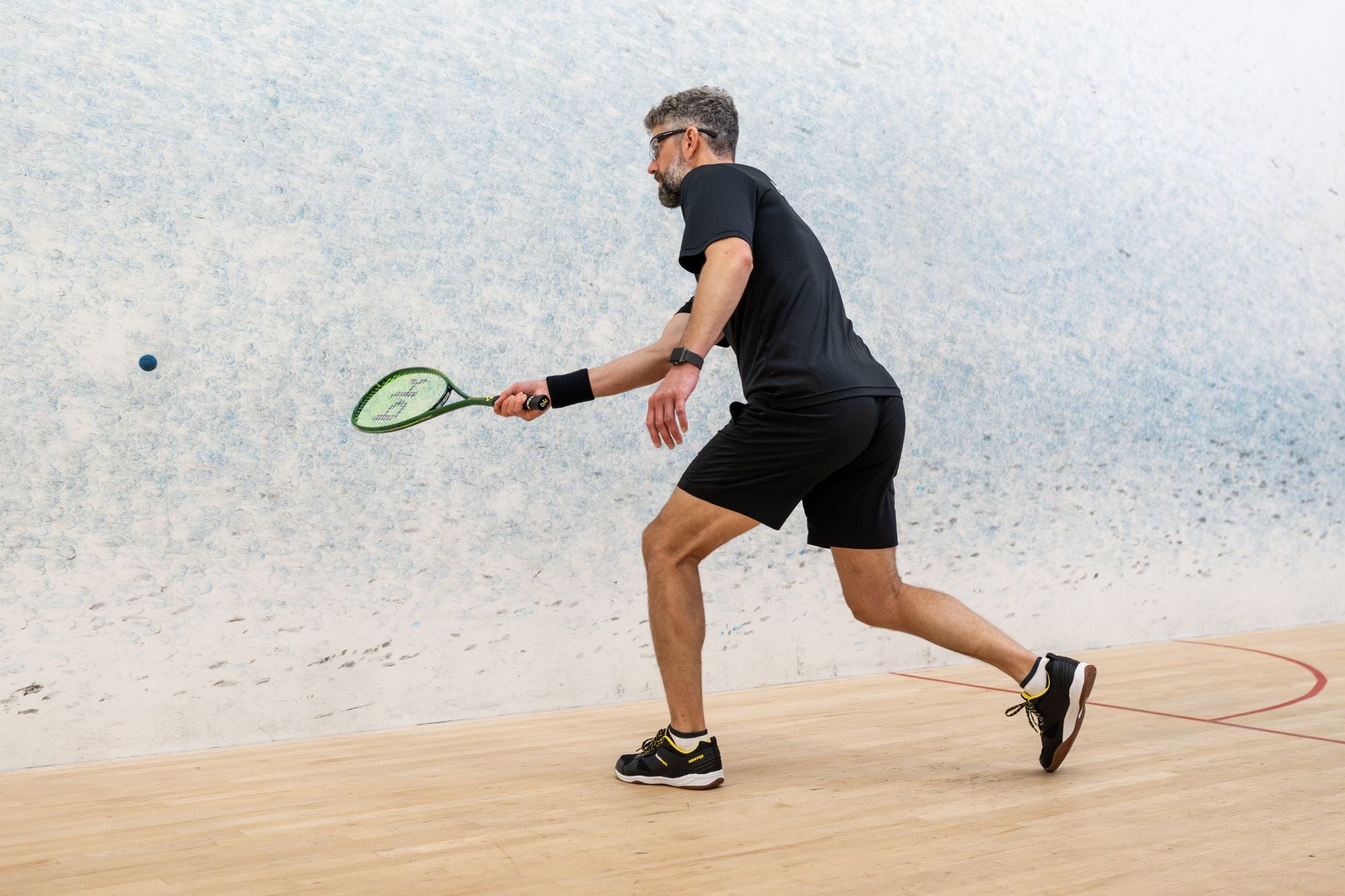 Man playing squash on a court with a light-colored wall and wooden floor.