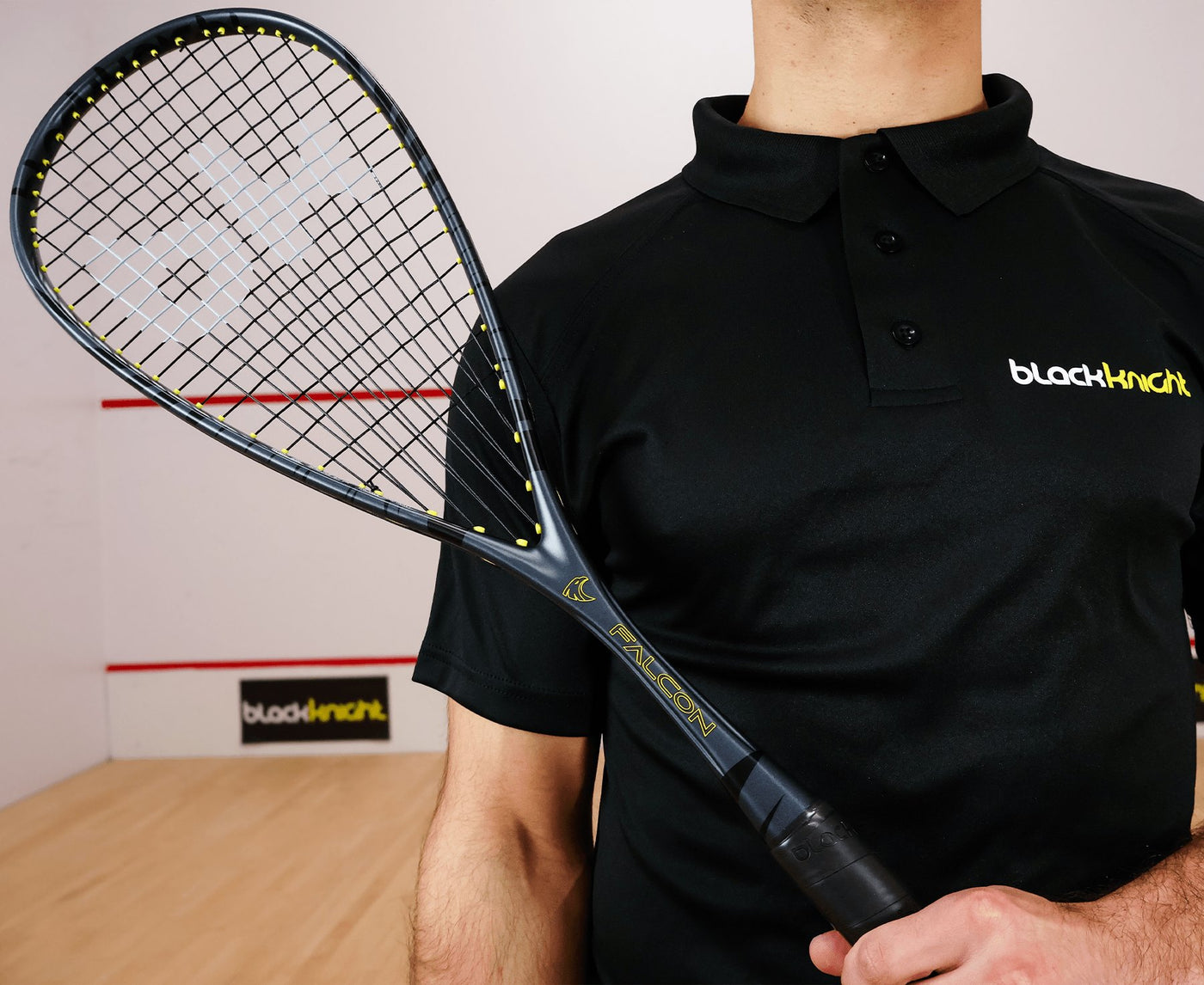 Person holding a squash racket with a black shirt featuring 'blacknight' branding in a squash court.