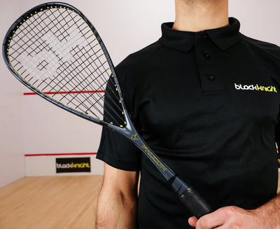 Person holding a squash racket with a black shirt featuring 'blacknight' branding in a squash court.