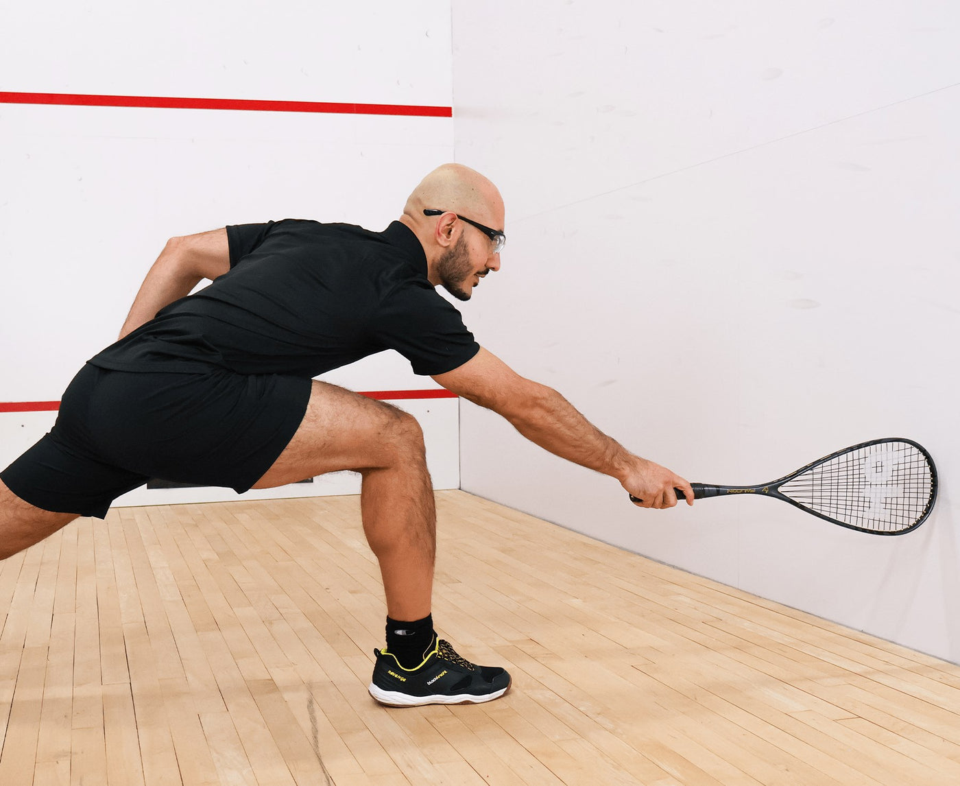 Man playing squash on a court with a white wall and wooden floor.